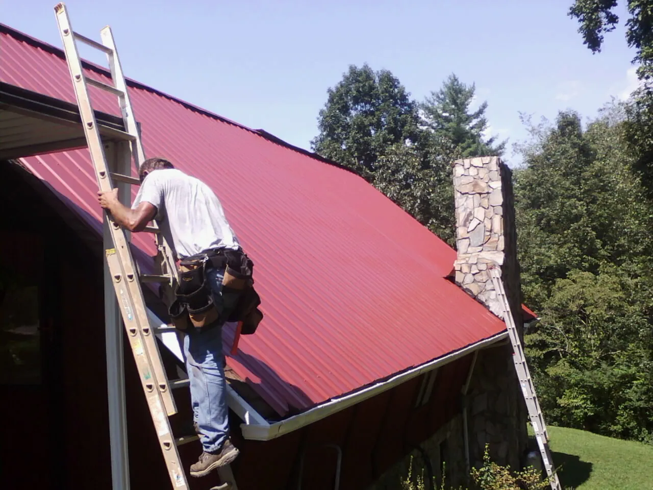 Roofer on a ladder beside a red metal roof and stone chimney — Soffit & Fascia Repair by Roofing Brothers in Petros